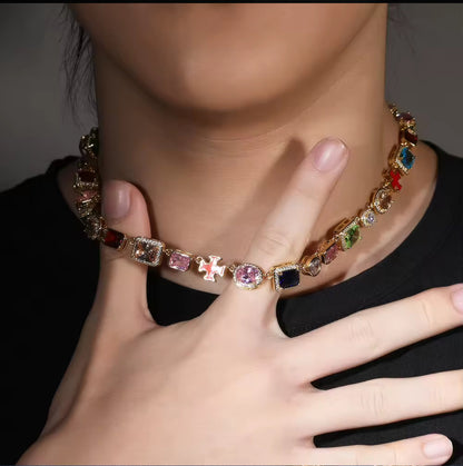 A person wearing a gold-plated chain necklace adorned with colorful gemstones and a cross pendant.
